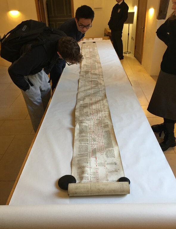 Students in a university library examining a long historical manuscript scroll laid out on a table