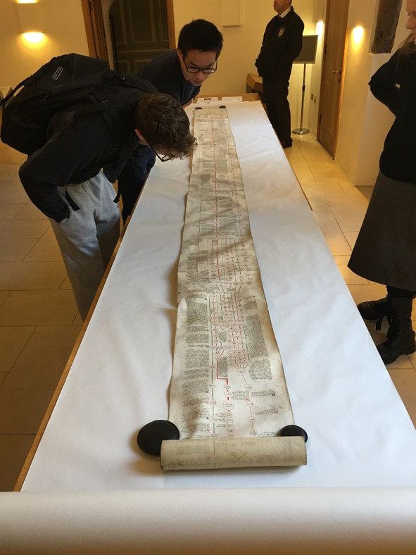 Students in a university library examining a long historical manuscript scroll laid out on a table