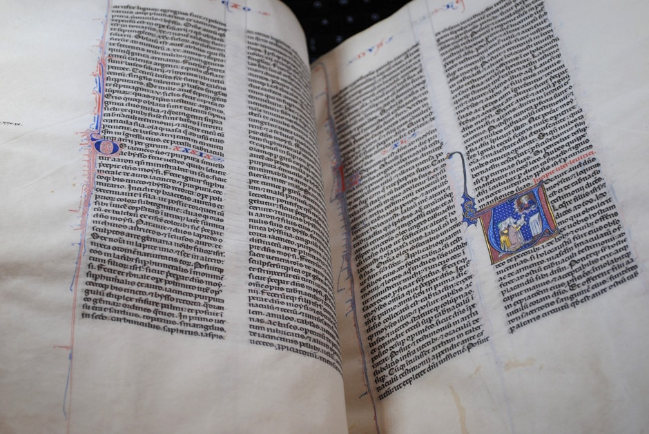 Open illuminated medieval manuscript showing Latin text in two columns with decorative initials and a small painted miniature