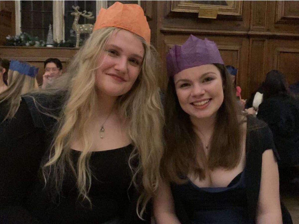 Two people wearing formal attire indoors, smiling at the camera, wearing Christmas paper hats