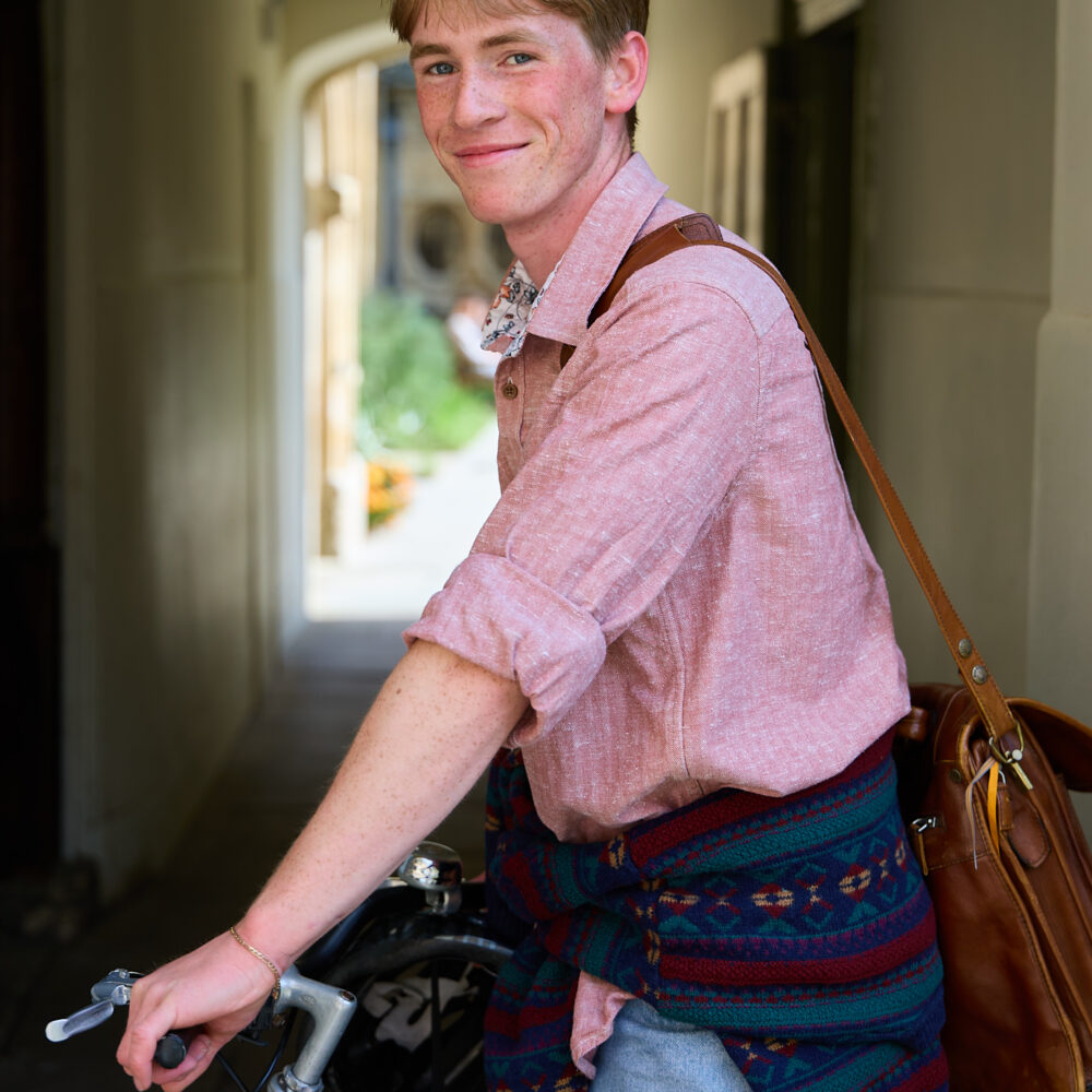 Person standing with a bicycle wearing a leather satchel, smiling slightly