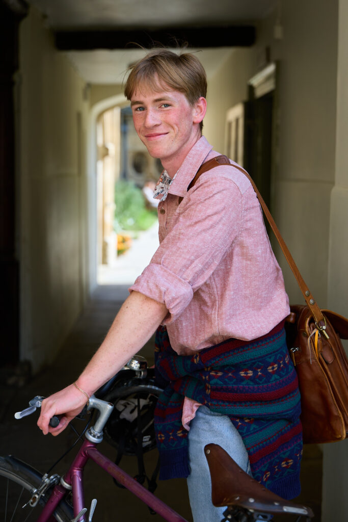Person standing with a bicycle wearing a leather satchel, smiling slightly