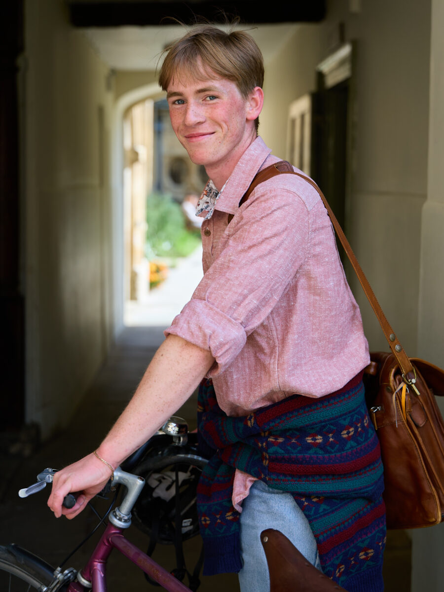 Person standing with a bicycle wearing a leather satchel, smiling slightly