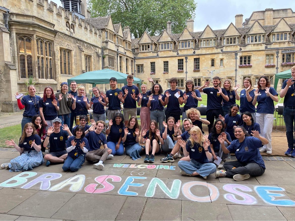 Large group of people sitting together outdoors on the quad with a chalk drawn writing 'Welcome to Brasenose'