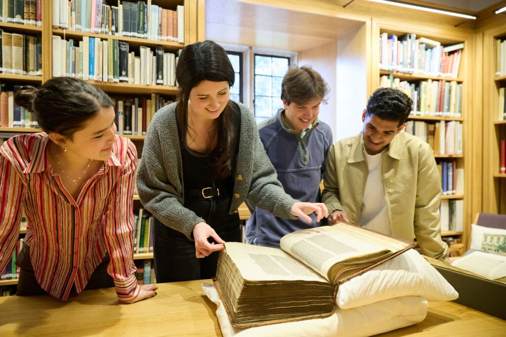 A librarian and three students examining a large, rare book on a table in a library