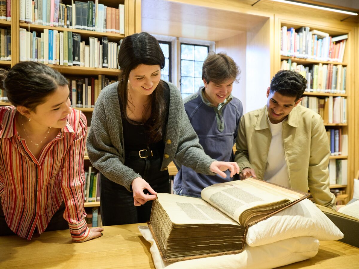 A librarian and three students examining a large, rare book on a table in a library