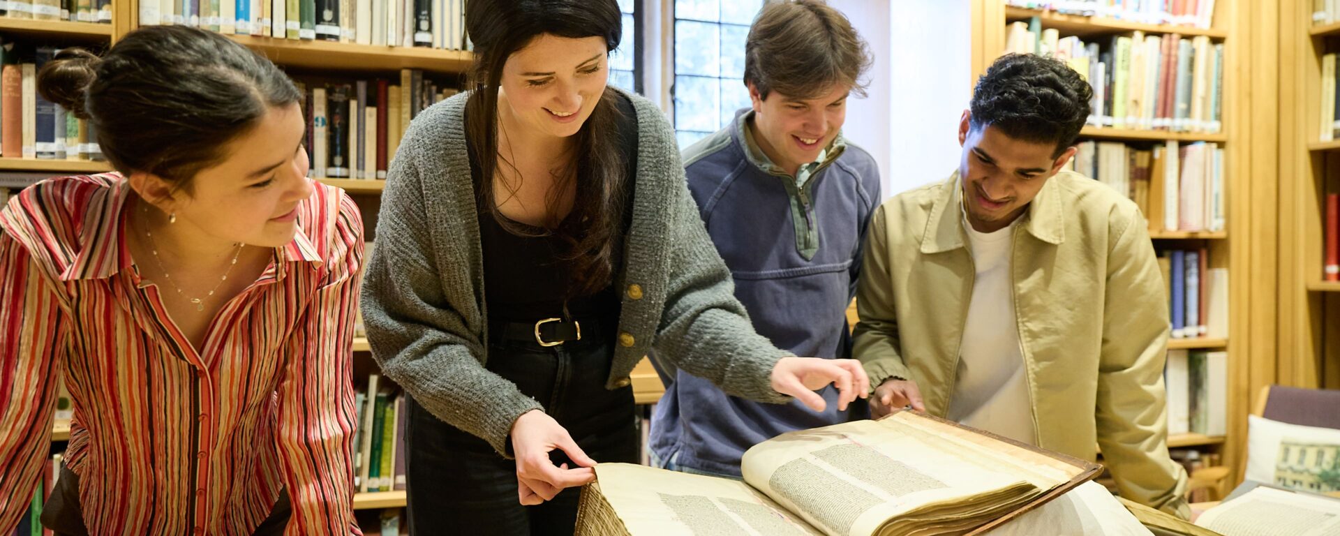 A librarian and three students examining a large, rare book on a table in a library