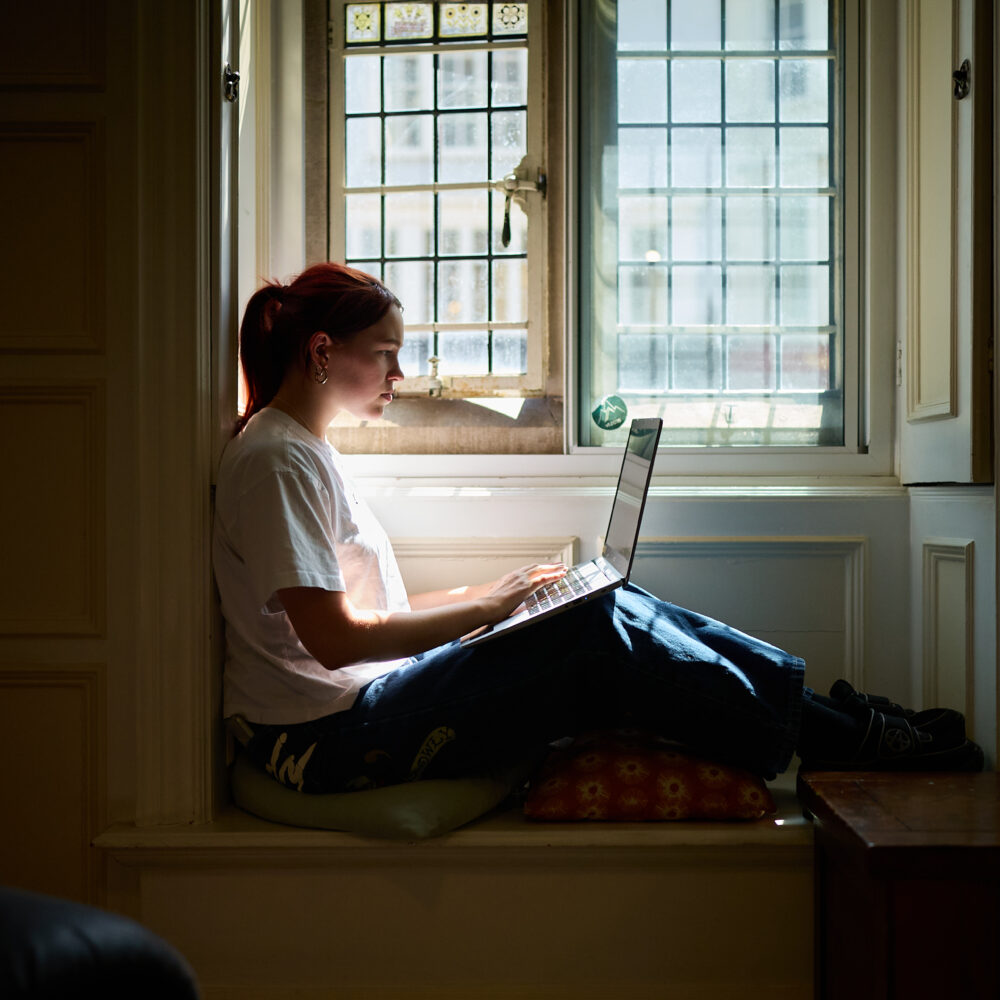 Person sitting by a window in an alcove, working on a laptop
