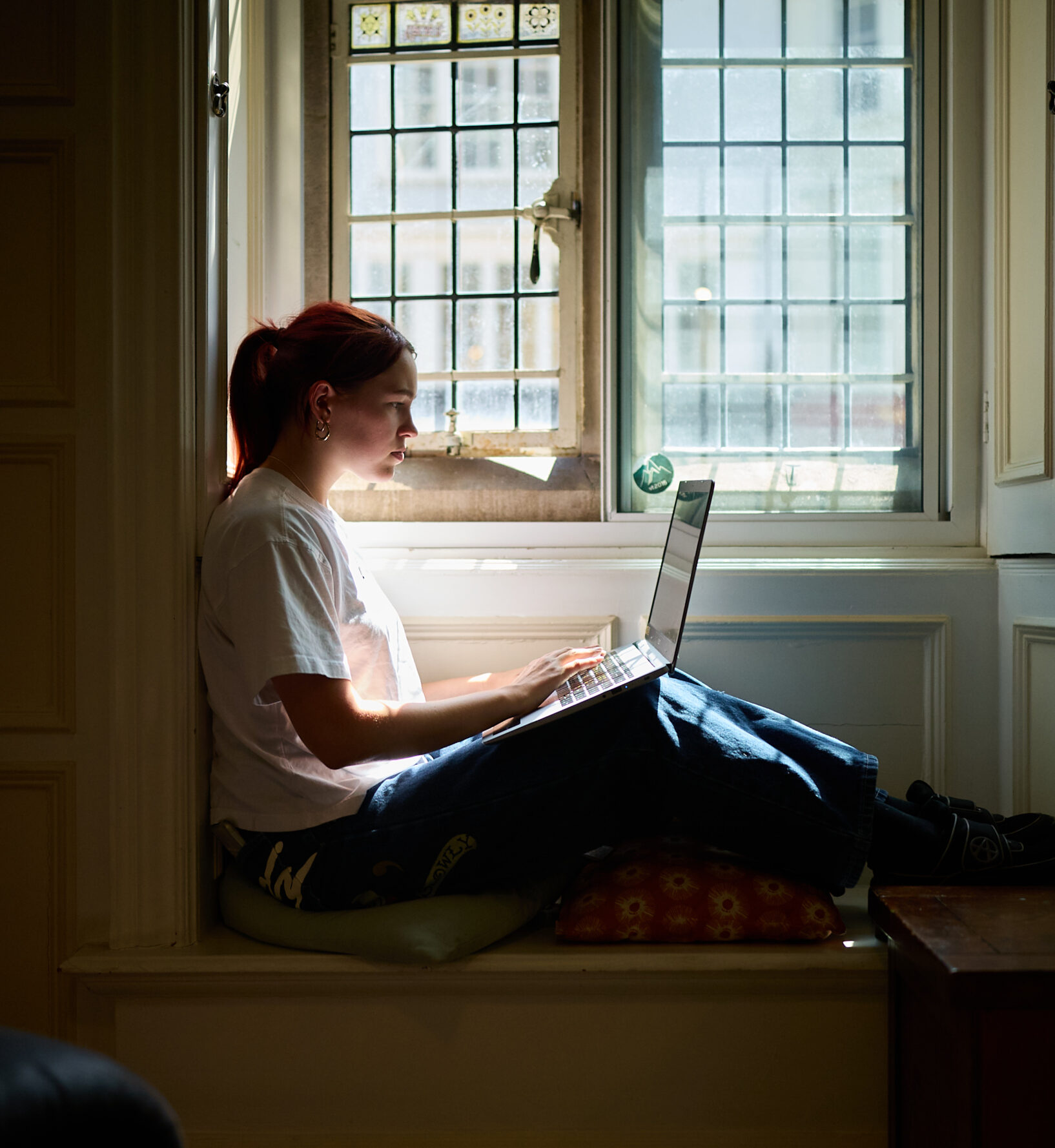 Person sitting by a window in an alcove, working on a laptop