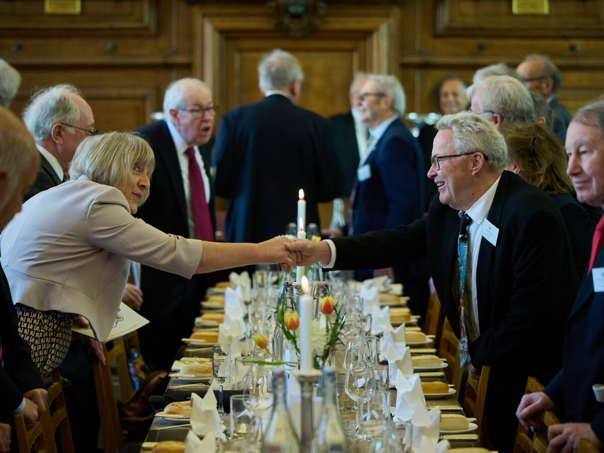Formal dinner table set indoors, with Jubilee Lunch guests standing and talking nearby.