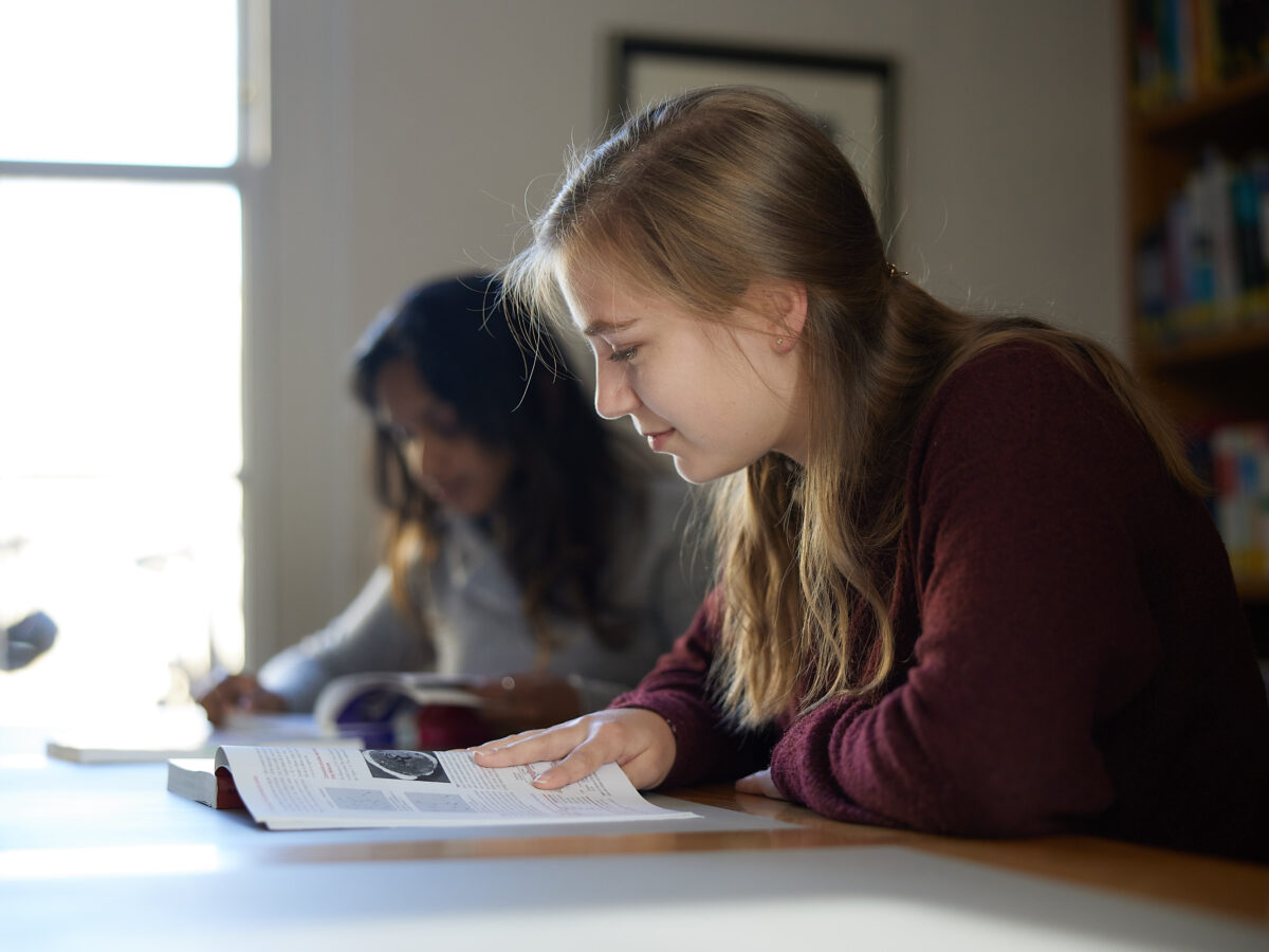 Two people seated indoors at a desk, looking down at papers or a notebook