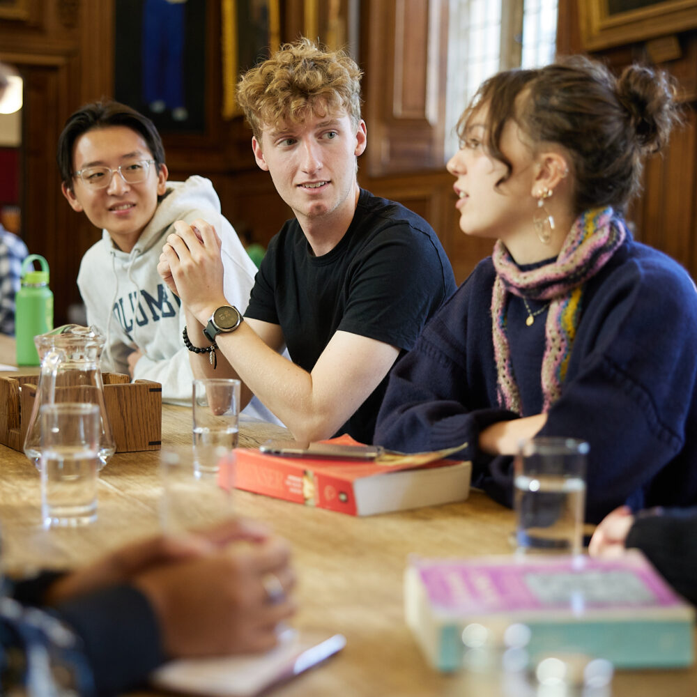 Small group of people sitting around a table indoors, talking with books in front of them