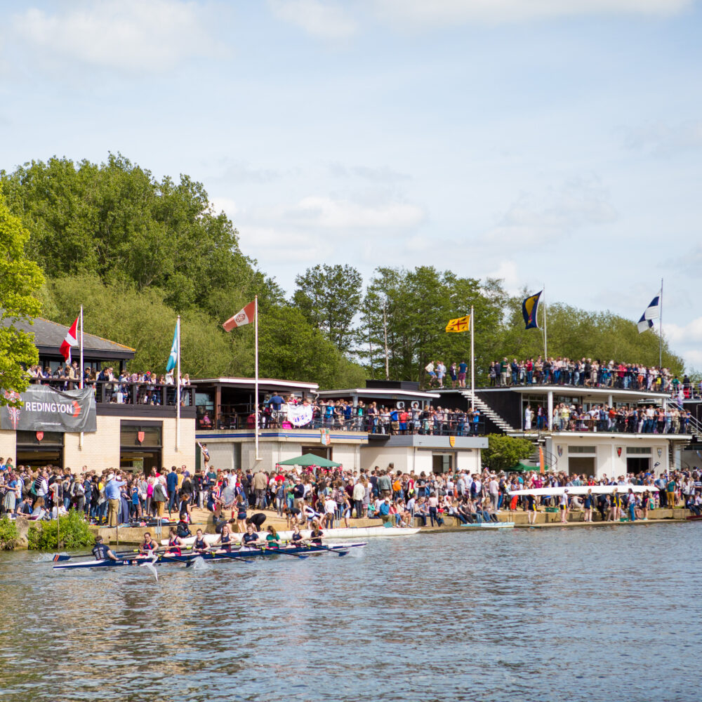 Eights on a river with spectators gathered along the banks on a sunny day