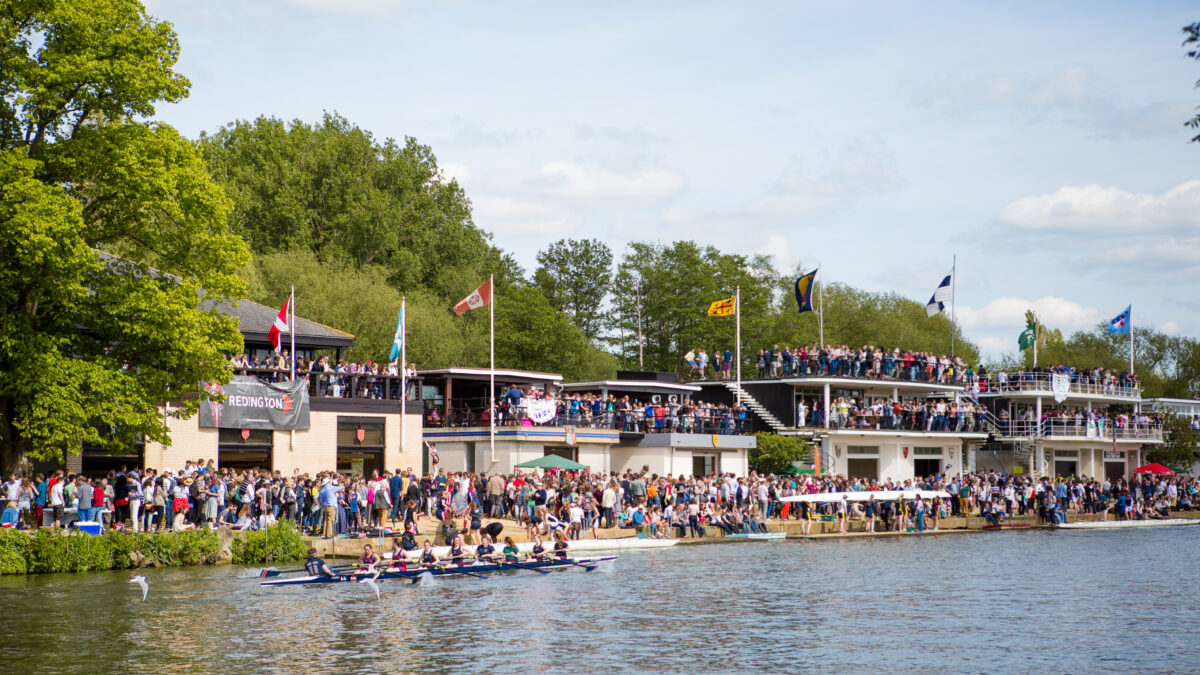 Eights on a river with spectators gathered along the banks on a sunny day