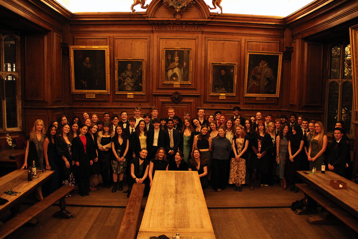 Students gathered together for a group photo at a formal dinner in a wood-panelled college room
