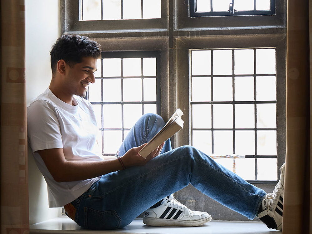 Student sitting in a window alcove reading a book in a college room