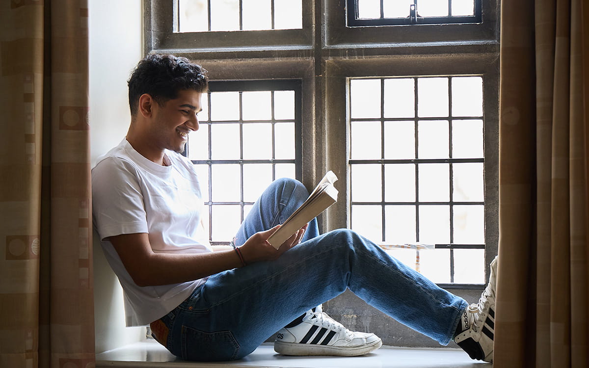 Student sitting in a window alcove reading a book in a college room