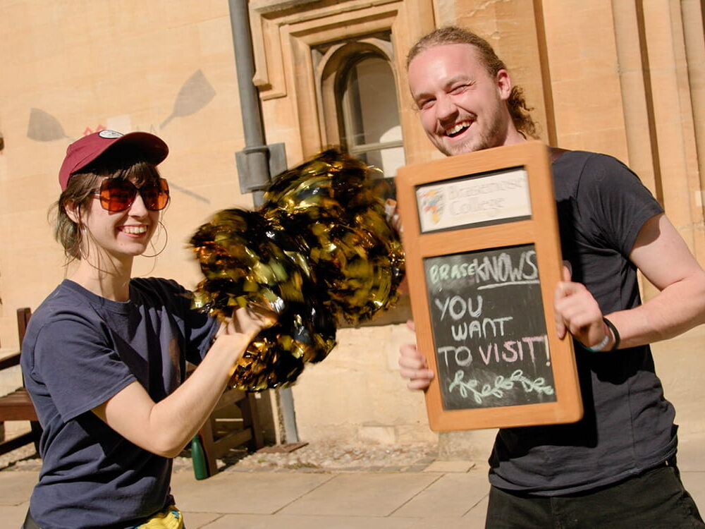 Student volunteers welcoming visitors, holding a sign reading ‘Brasenose – you want to visit here