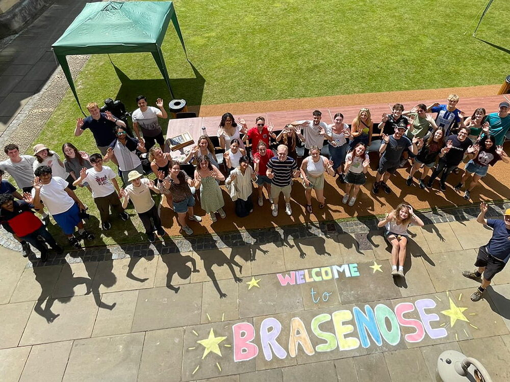 A group of people posing for a photograph taken from above with 'Welcome to Brasenose' chalked on the stone paving in front of them