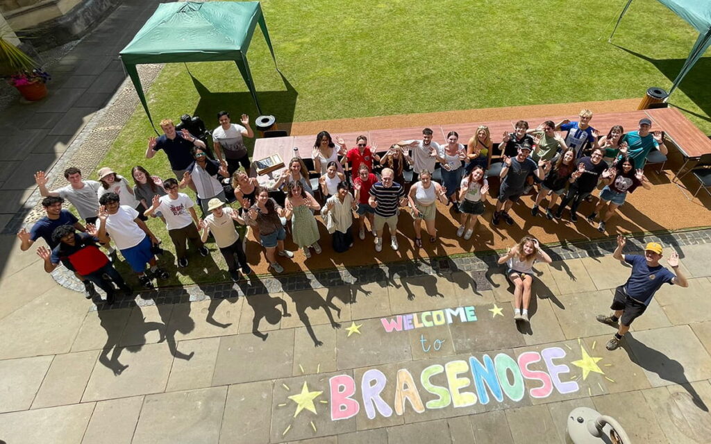 A group of people posing for a photograph taken from above with 'Welcome to Brasenose' chalked on the stone paving in front of them