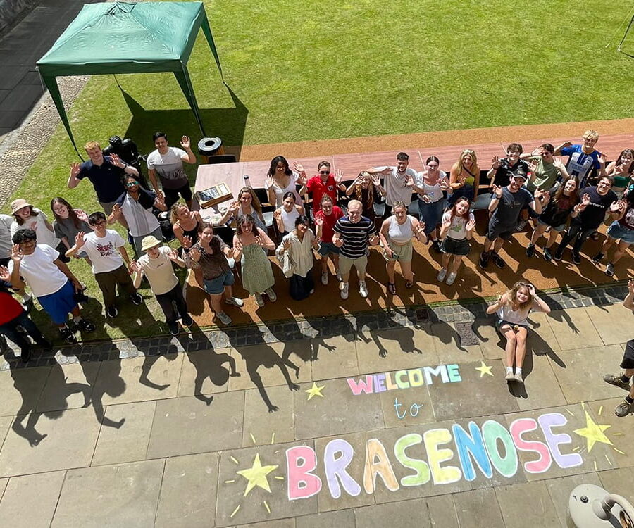 A group of people posing for a photograph taken from above with 'Welcome to Brasenose' chalked on the stone paving in front of them