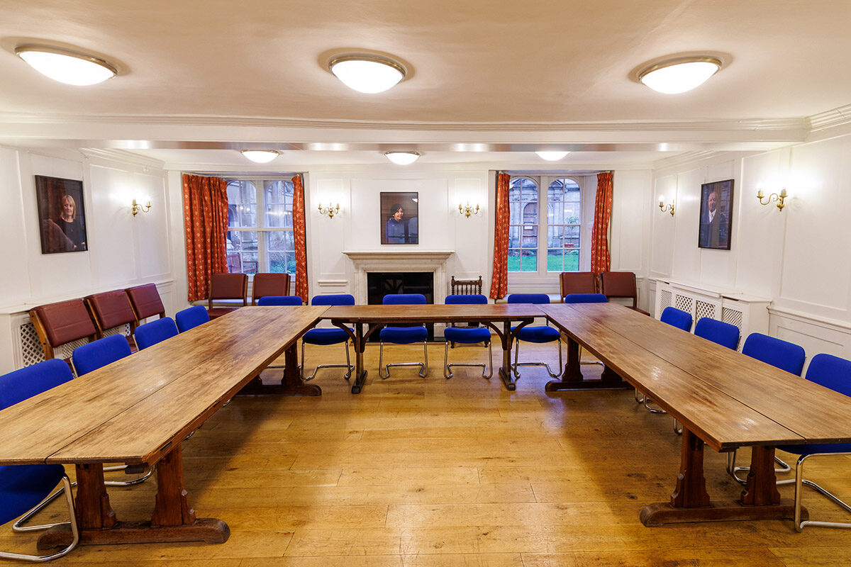 A formal meeting room at Brasenose College with long wooden tables arranged in a U-shape, blue chairs, white panelled walls, portraits, and a central fireplace beneath large windows.