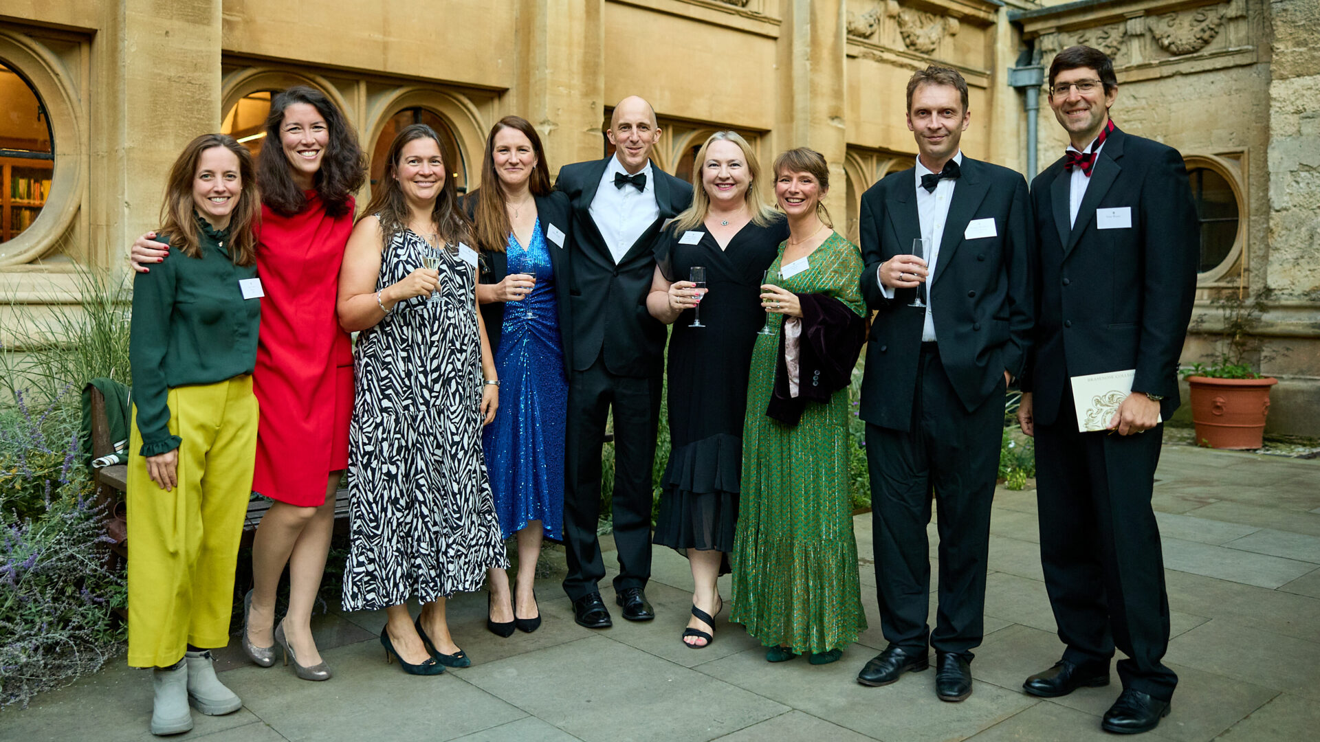 a group of nine alumni dressed in black tie standing in deer park quad