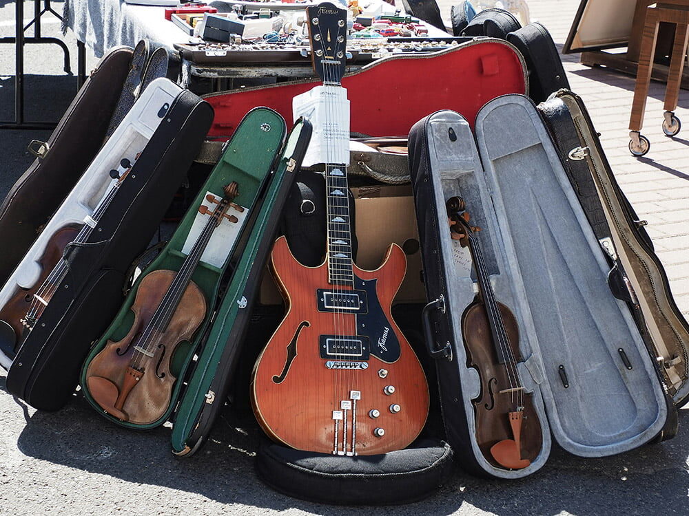 A collection of musical instruments in open cases, including violins and an electric guitar, arranged outdoors