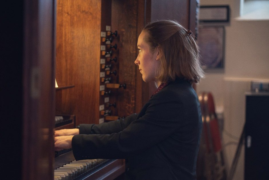 Lucy Morrell playing an organ, viewed from the side, with her hands on the keyboards and organ stops visible beside her.