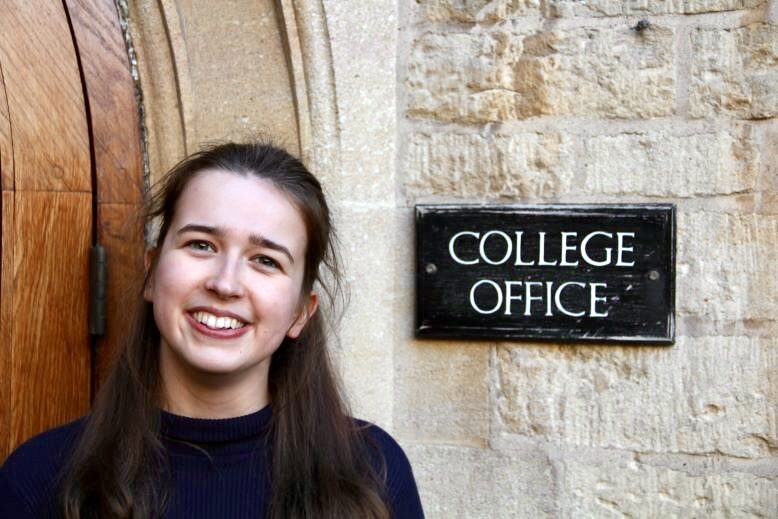 Photo of Holly smiling, stood outside the College Office door sign