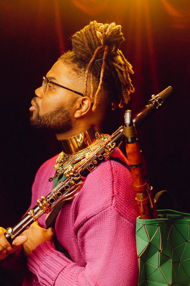 Musician in profile wearing a bright pink jumper, holding a clarinet, with a bassoon strapped across his back